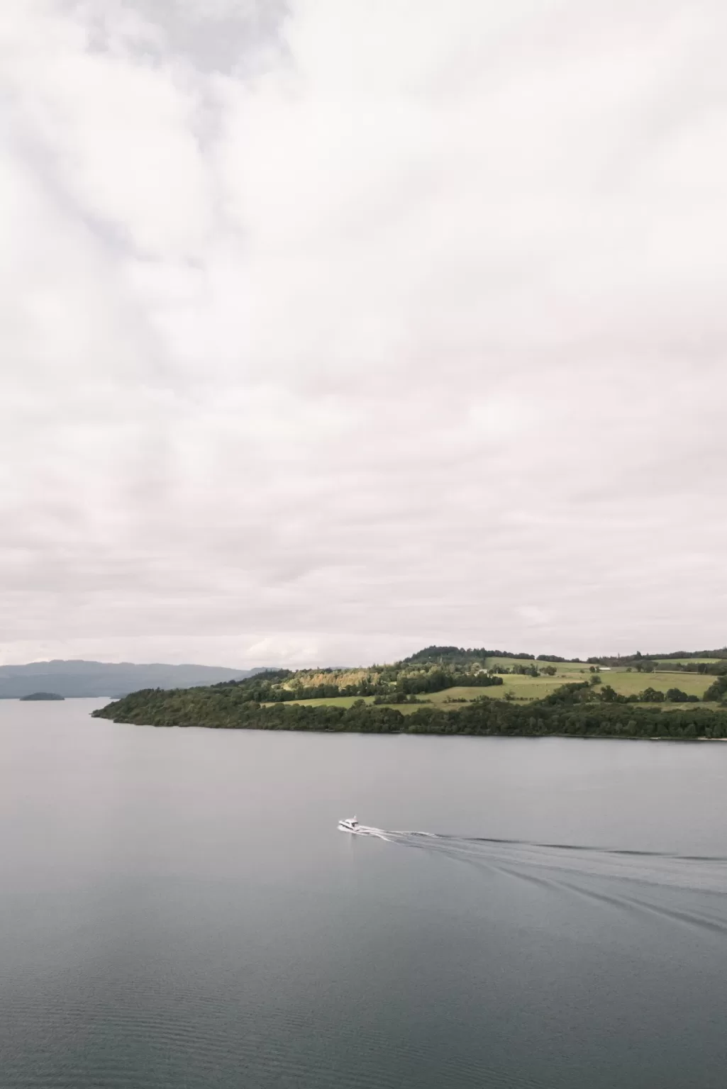 Small boat crossing Loch Lomond near Boturich Castle under a soft, overcast sky.