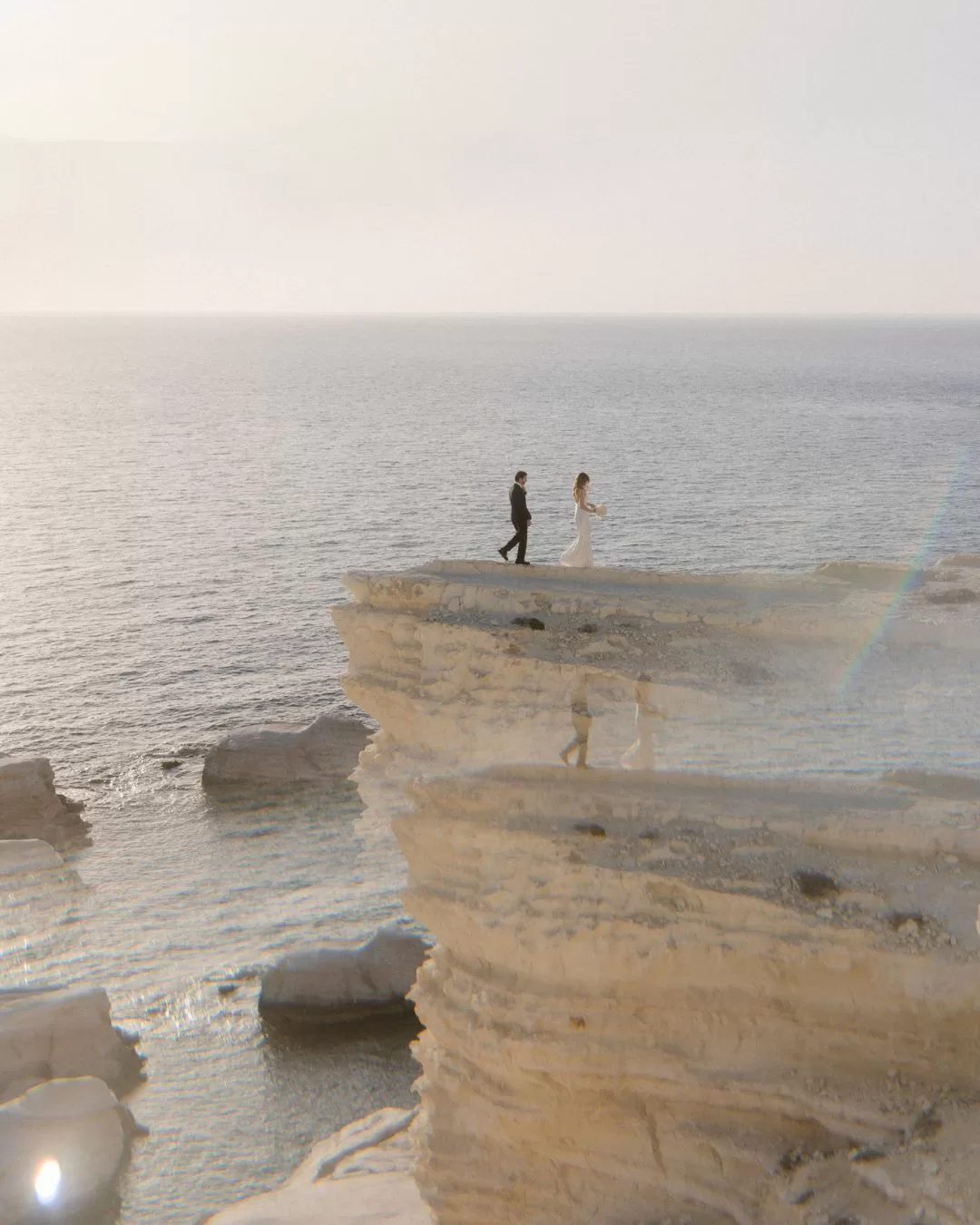A newlywed couple stands on a rocky cliff overlooking the Mediterranean Sea, captured by Karina Leonenko