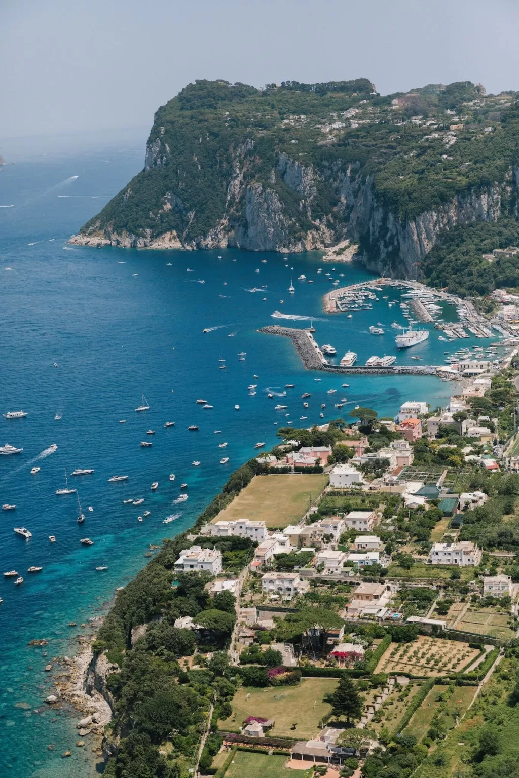 Aerial view of Capri’s dramatic coastline and marina, showing the turquoise sea dotted with boats and lush greenery.