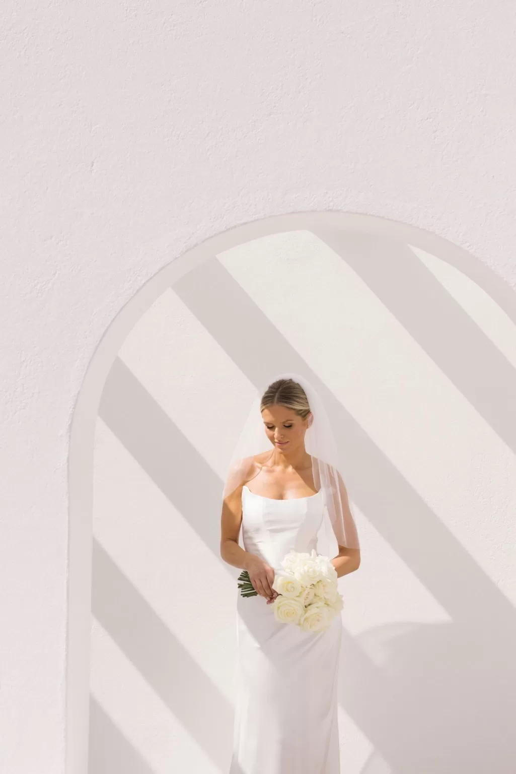 Bride standing beneath a sunlit archway at Casa Arte, holding a bouquet of white roses against the venue’s clean white walls.