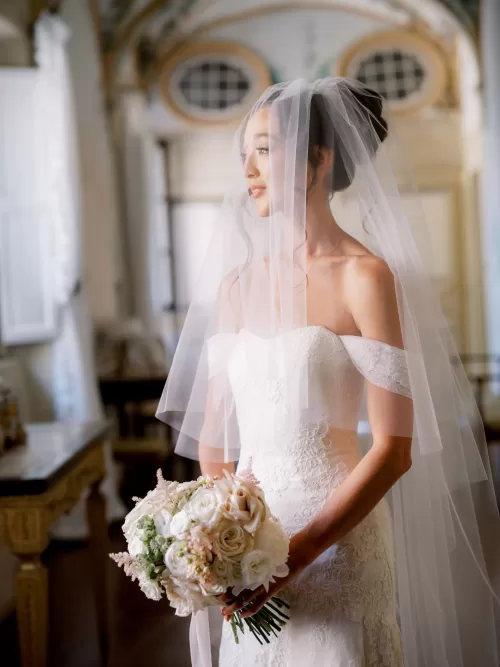 Bride in lace gown holding bouquet inside the historic interiors of Castello di Celsa.