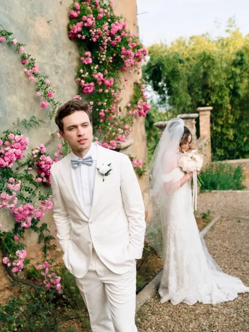 Groom in cream suit in the foreground while bride in veil holds bouquet beside rose-covered wall at Castello di Celsa.