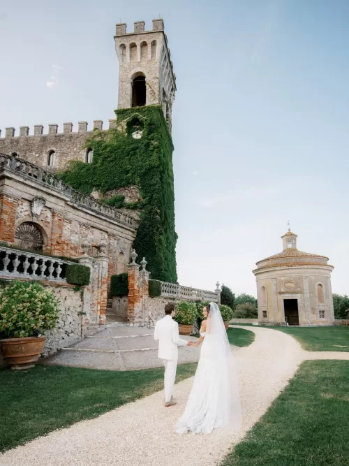 Bride and groom walking along the gravel path between the castle façade and round chapel at Castello di Celsa.