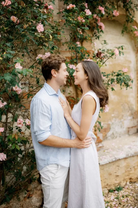 Couple embracing beneath soft pink roses climbing the stone walls of Castello di Celsa.