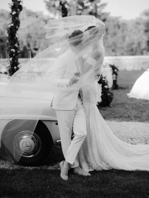 Romantic black and white portrait of the couple beside a vintage car at Castello di Celsa, bride’s veil flowing in the breeze.