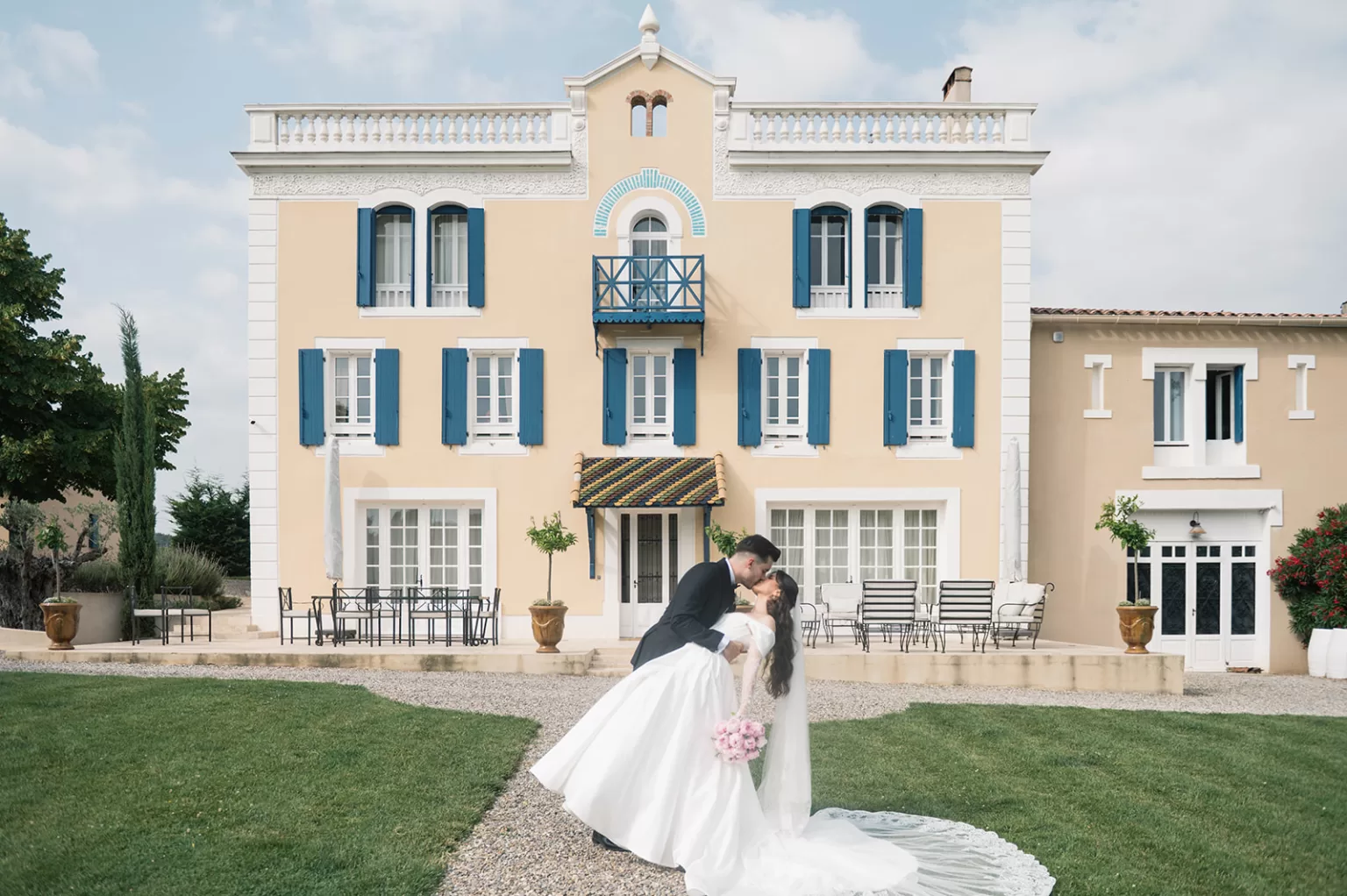 Bride and groom sharing a kiss in front of the Chateau Canet facade with blue shutters and formal gardens.