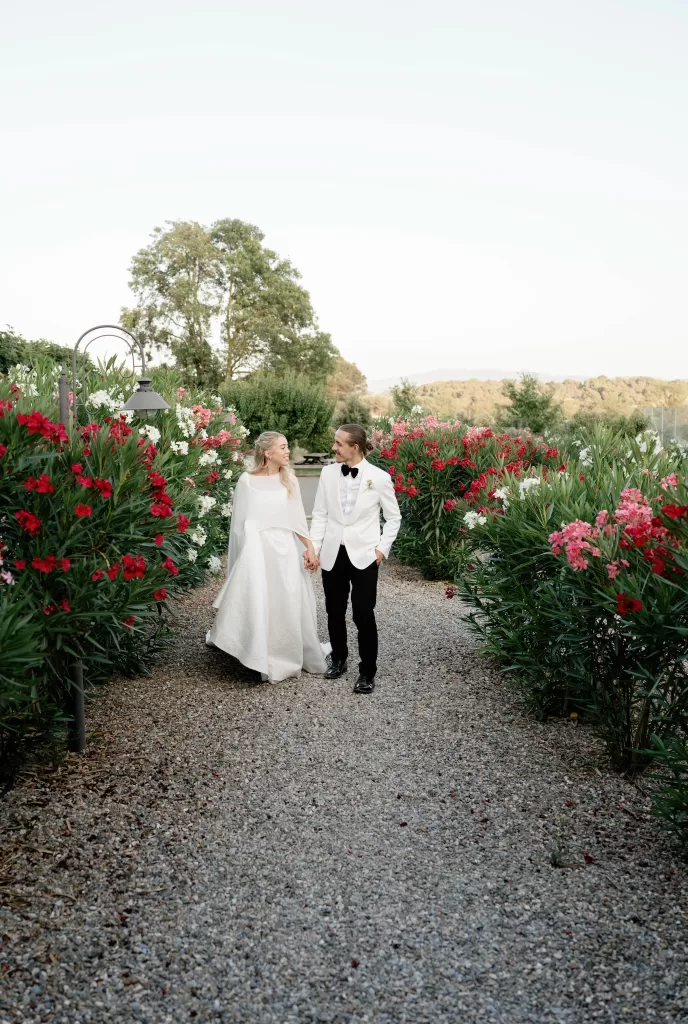 Bride and groom walking hand in hand through vibrant flowering gardens at Château Canet, surrounded by red and pink blooms under soft evening light.