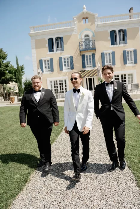 Groom in a white tuxedo jacket walking with groomsmen in black suits across the Château Canet lawn, the château façade creating a refined backdrop.