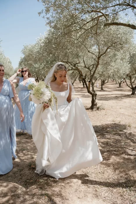 Bride walking through the olive grove at Château Canet with bridesmaids in soft blue dresses, carrying a white bouquet beneath the Mediterranean sun.