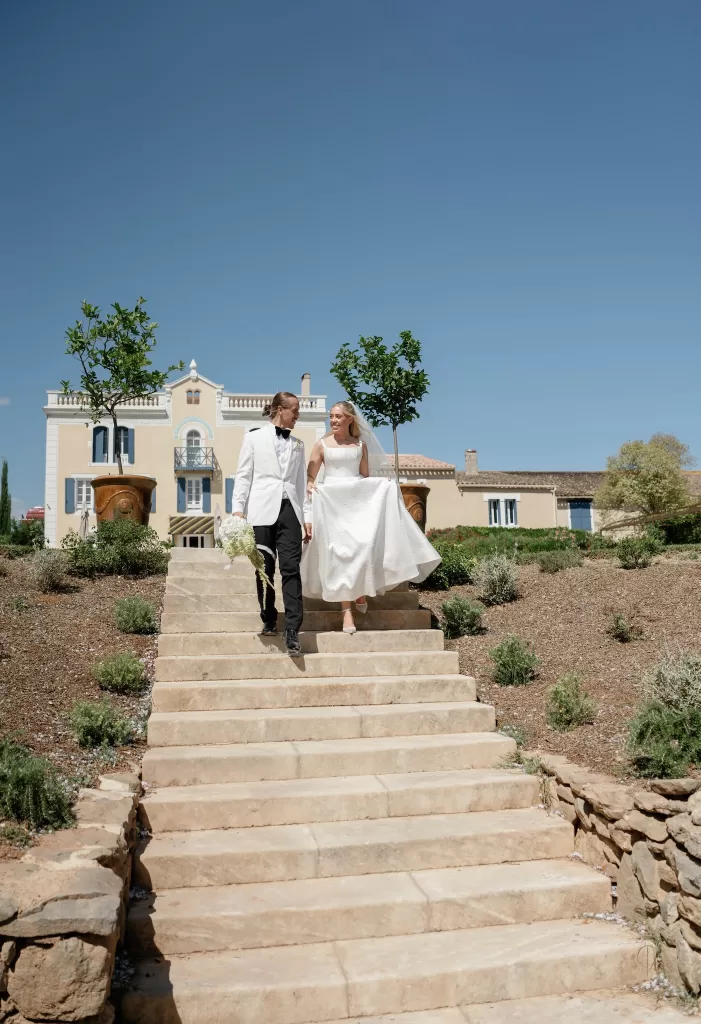Bride and groom descending the grand stone staircase at Château Canet, with the elegant château façade and clear blue sky creating a timeless South of France backdrop.