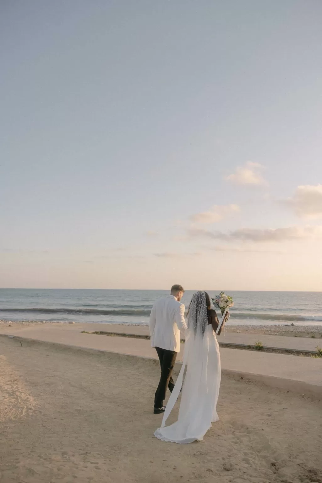Bride and groom walking along the sandy beach at Coral Residences under a soft golden sunset.