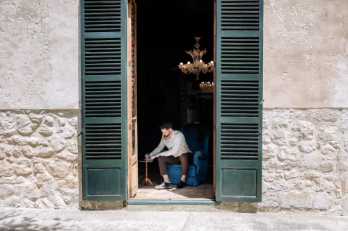 Groom writing his vows inside Finca Morneta, seated by open green shutters with chandeliers glowing behind him.