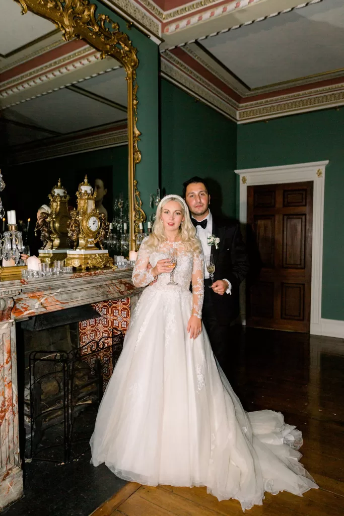 Newlyweds standing by an ornate fireplace inside Garthmyl Hall, surrounded by rich green walls, gilded mirrors and candlelight.