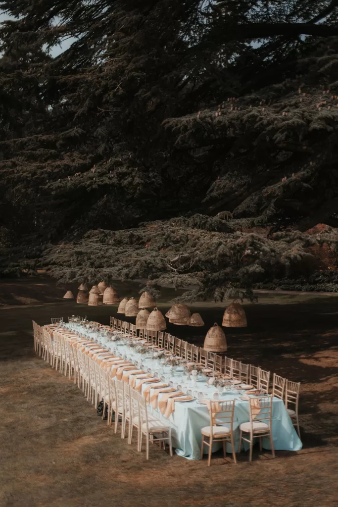 Long outdoor reception table set beside the river at Garthmyl Hall, with woven pendant lights overhead and soft pastel table styling at dusk.