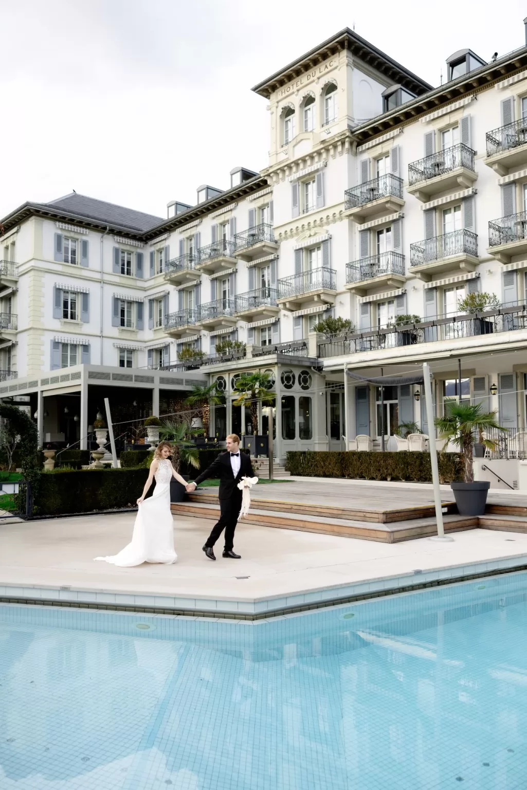 Bride and groom walking hand in hand beside the pool at Grand Hotel du Lac, framed by the elegant historic facade.