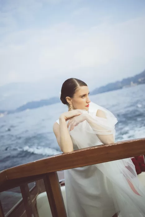 Bride leaning on wooden boat railing on Lake Como, veil flowing softly in the breeze near Hotel Ristorante La Darsena.