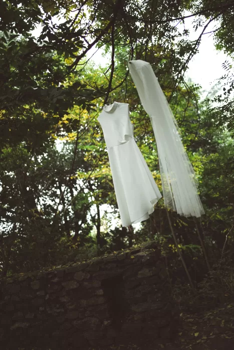 Minimalist white wedding dress and veil hanging from tree branches above stone wall at Hotel Ristorante La Darsena.