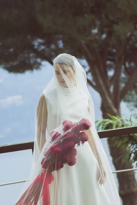Bride holding deep red bouquet beneath sheer veil on terrace at Hotel Ristorante La Darsena with Lake Como backdrop.