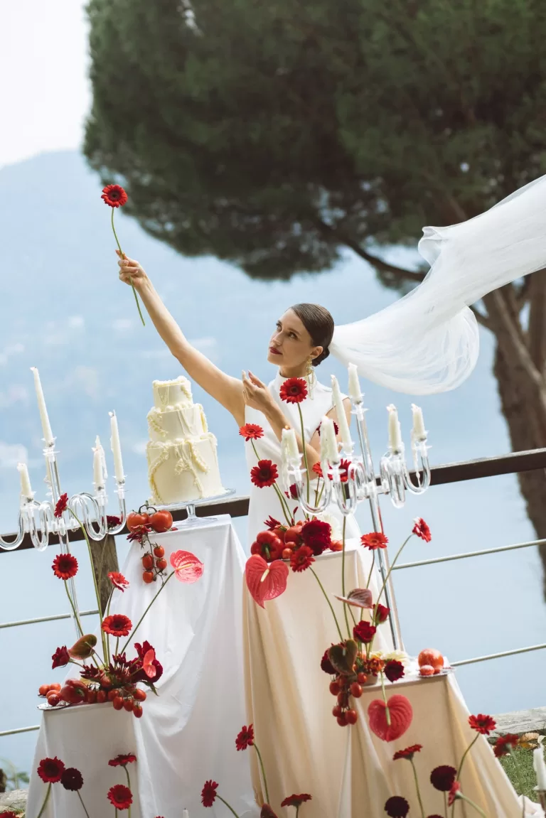 Bride lifting a single red bloom beside tiered wedding cake on terrace at Hotel Ristorante La Darsena overlooking the lake.