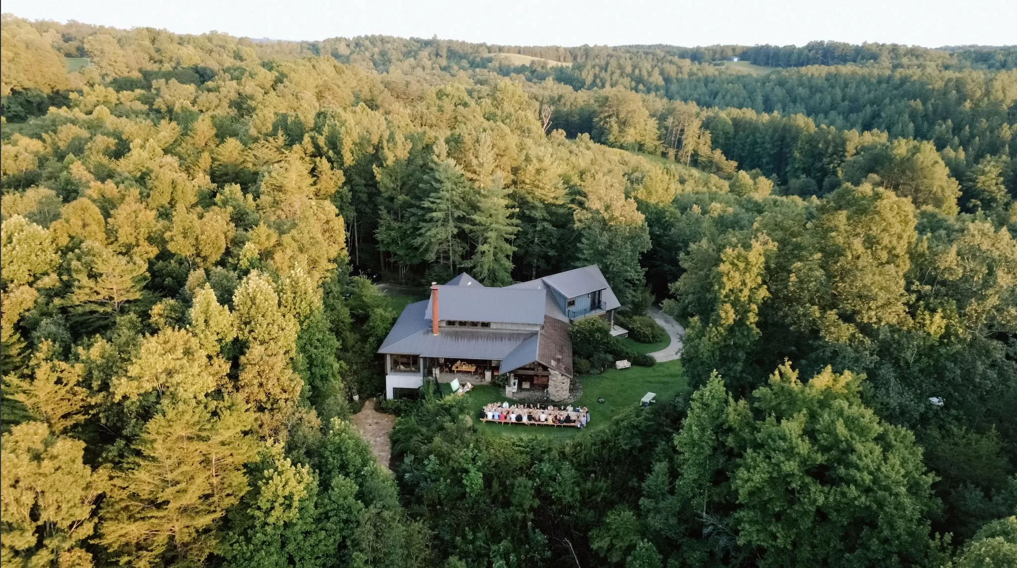 Aerial view of House Boheme surrounded by rolling forest and hills, with a long outdoor dinner table set on the lawn below.