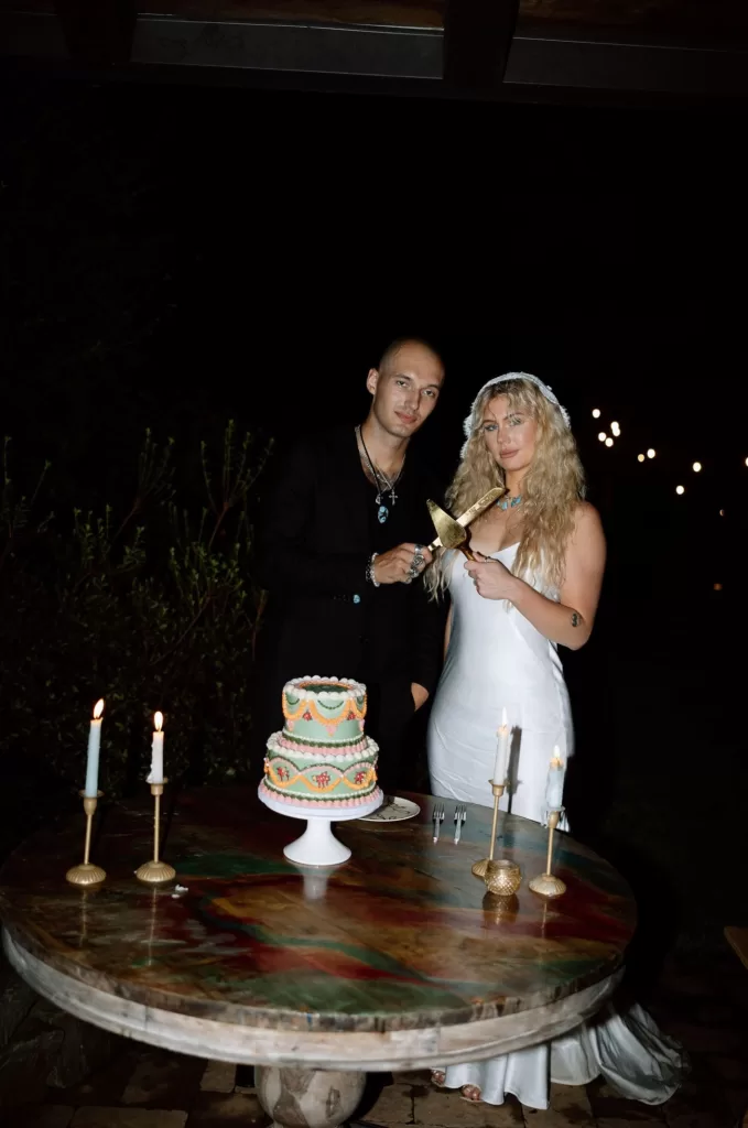 Couple cutting a decorated wedding cake at House Boheme by candlelight, standing beside a round marble table under the night sky.
