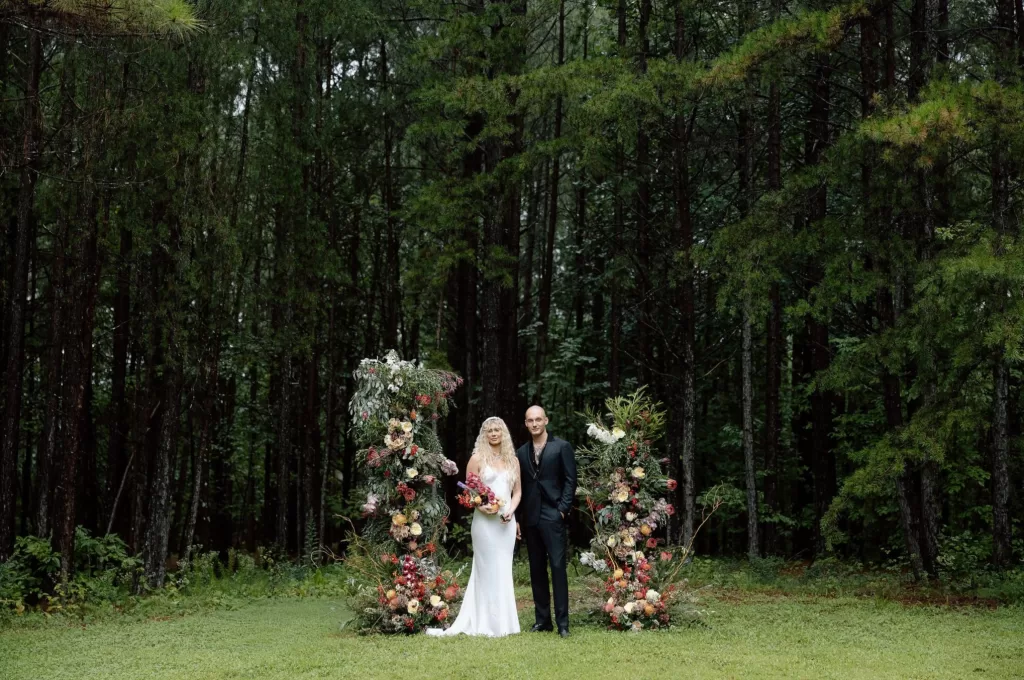 Bride and groom standing between two abundant floral arrangements in a forest clearing at House Boheme, surrounded by tall pine trees.