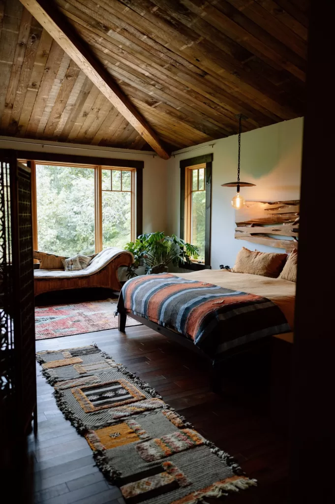 Rustic bedroom interior at House Boheme featuring wood beam ceilings, warm textures and large windows overlooking the surrounding forest.
