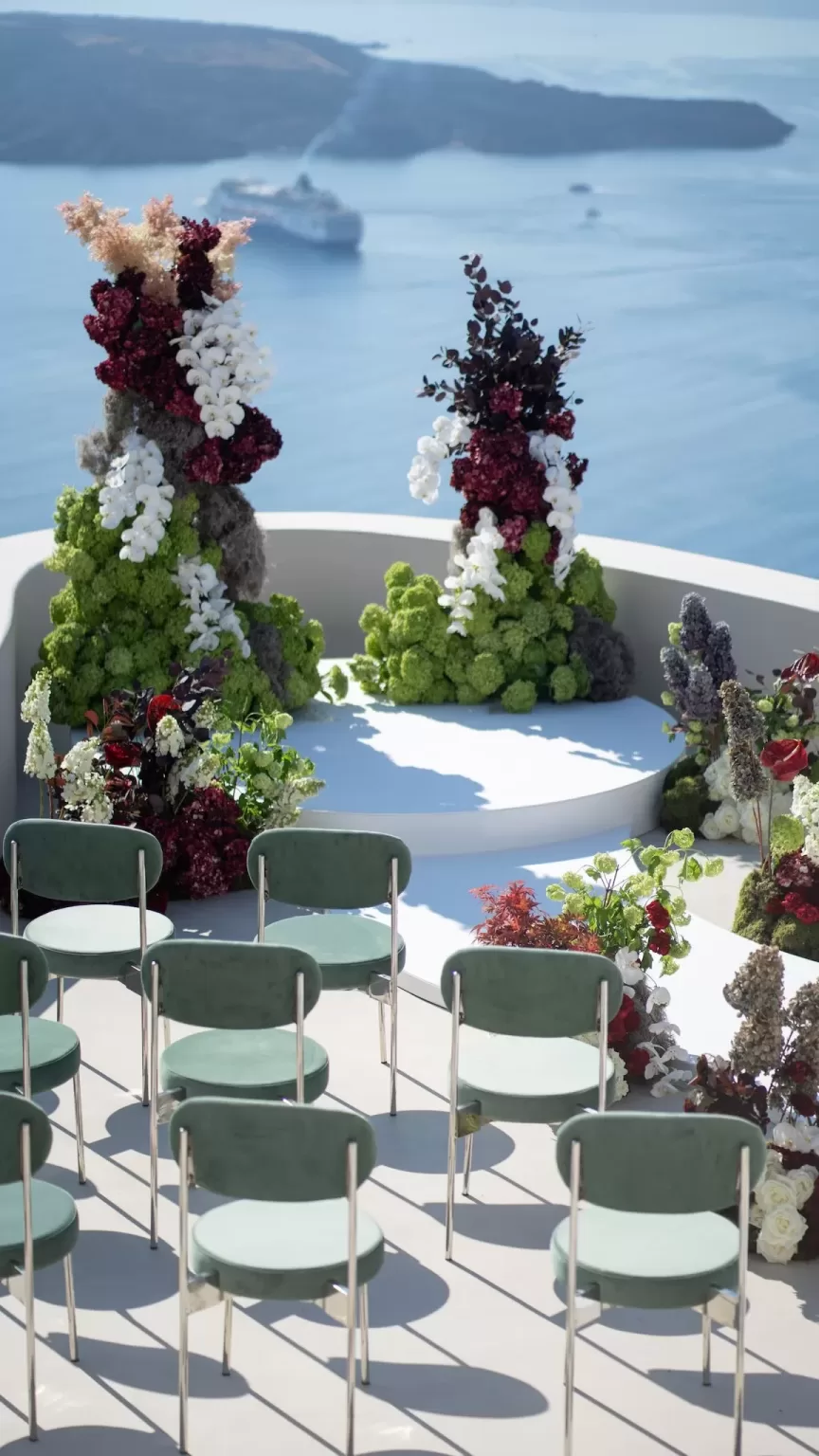 Modern wedding ceremony setup at Islet Santorini featuring sculptural floral installations and caldera views, with green velvet seating arranged around the altar.