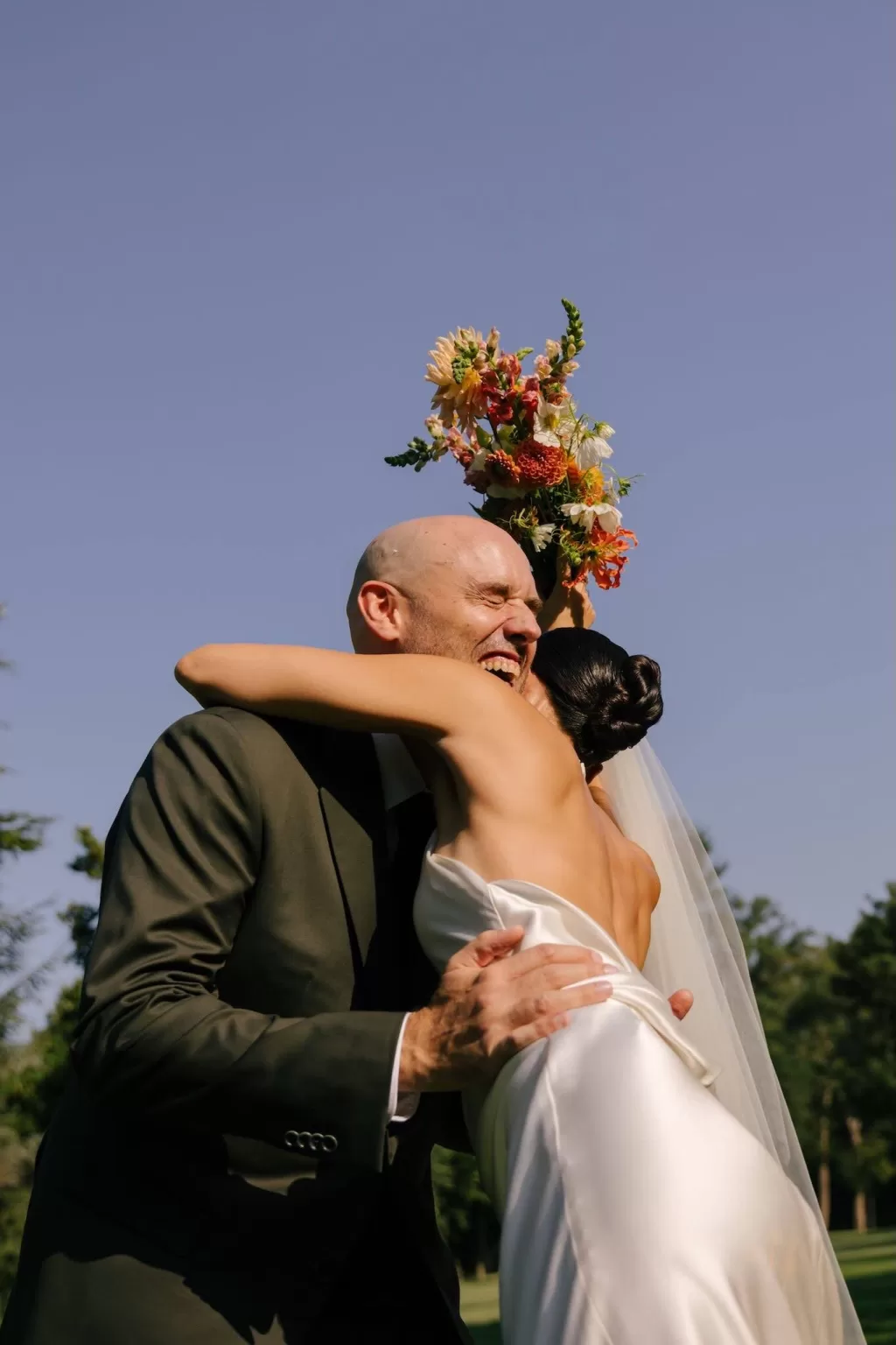 Groom embracing bride as she holds her bouquet aloft under a blue sky at La Farinera de Sant Lluis.