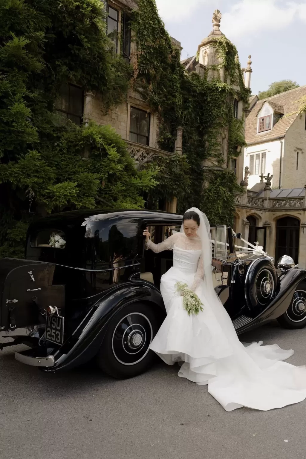 The bride steps gracefully from a classic black car at Manor House, holding her bouquet and veil.