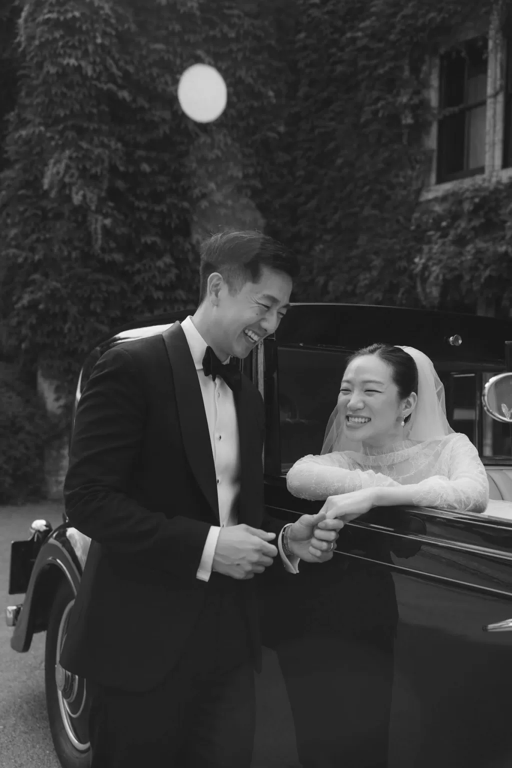 The couple share a joyful laugh beside their vintage car at Manor House, captured in timeless black and white.