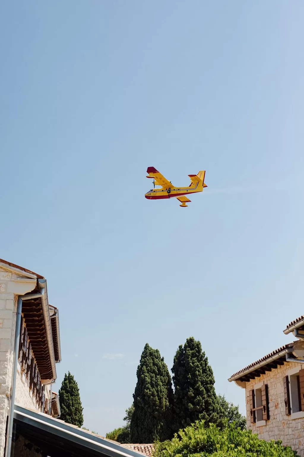 Bright yellow plane flying over Meneghetti Wine Hotel’s rustic stone buildings and cypress trees.