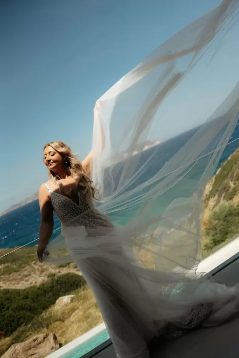 Bride lifting her flowing veil on sea-view balcony at Milos Cove with turquoise coastline behind.