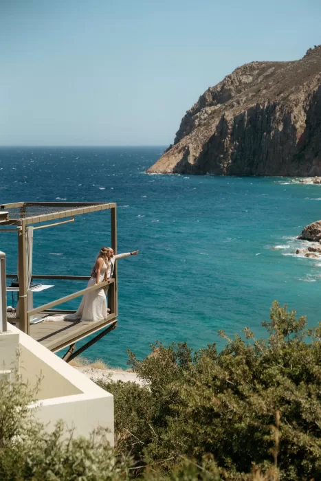 Bride and groom on cantilevered balcony at Milos Cove pointing towards the sparkling Aegean Sea.