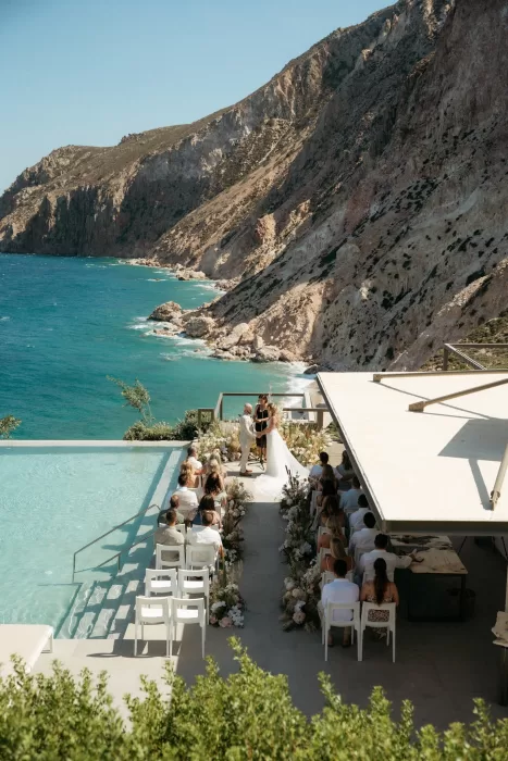 Wedding ceremony by the pool at Milos Cove with guests seated against dramatic seaside cliffs.