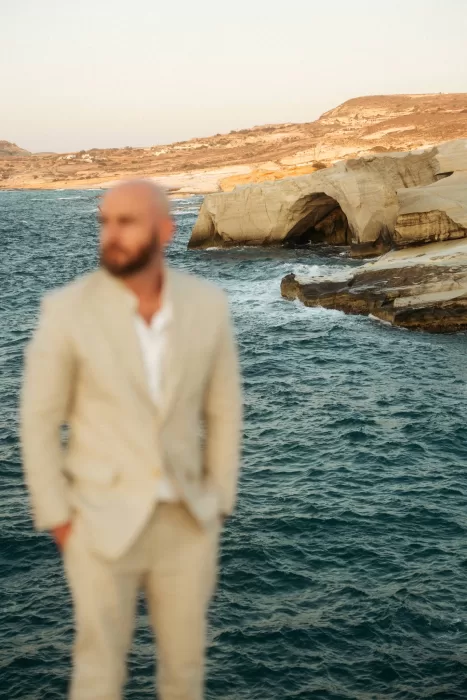 Groom standing by iconic rocky sea arch near Milos Cove with waves crashing below.