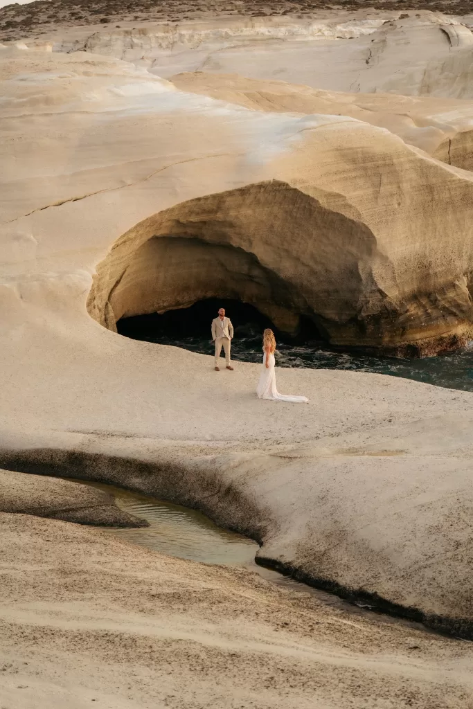 Bride and groom standing apart on dramatic rock landscape near Milos Cove sea cave.
