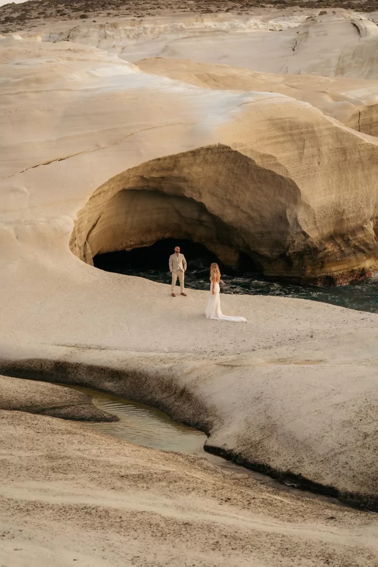 Bride and groom standing apart on dramatic rock landscape near Milos Cove sea cave.