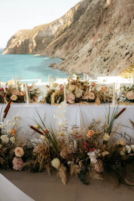 Seafront reception table at Milos Cove styled with abundant florals overlooking turquoise cliffs.