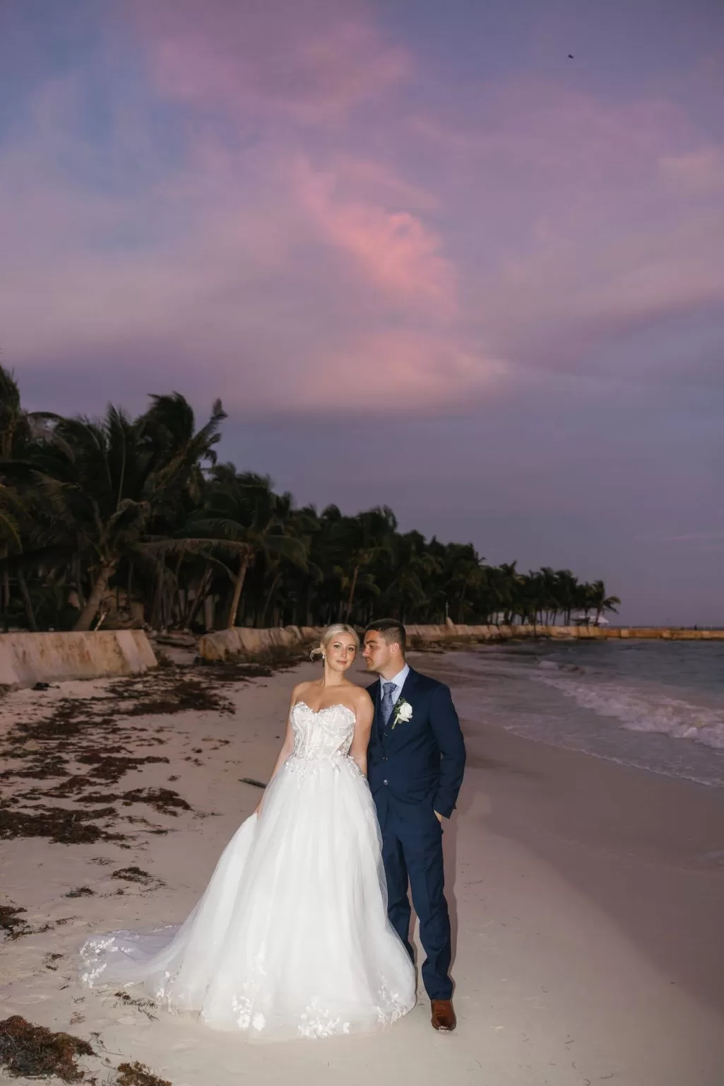 Newlyweds posing on the beach at sunset with palm trees and soft purple skies at Villa La Joya.