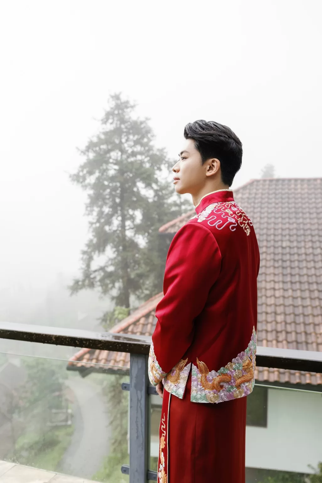 Groom in embroidered red traditional attire standing on a balcony at Ville de Mont Mountain Resort overlooking misty mountain scenery.