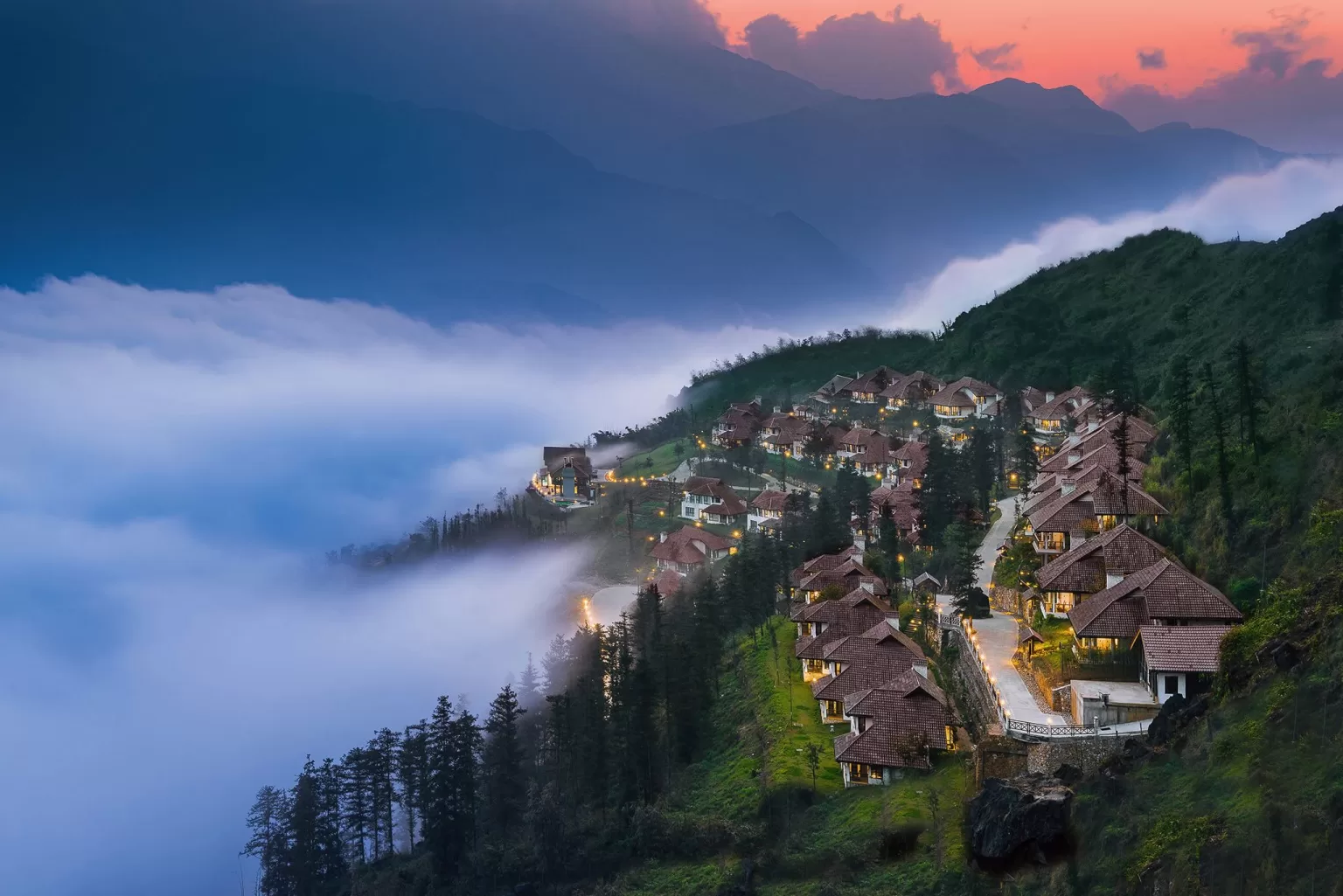 Hillside villas at Ville De Mont Mountain Resort illuminated at sunset above drifting clouds and forested slopes.
