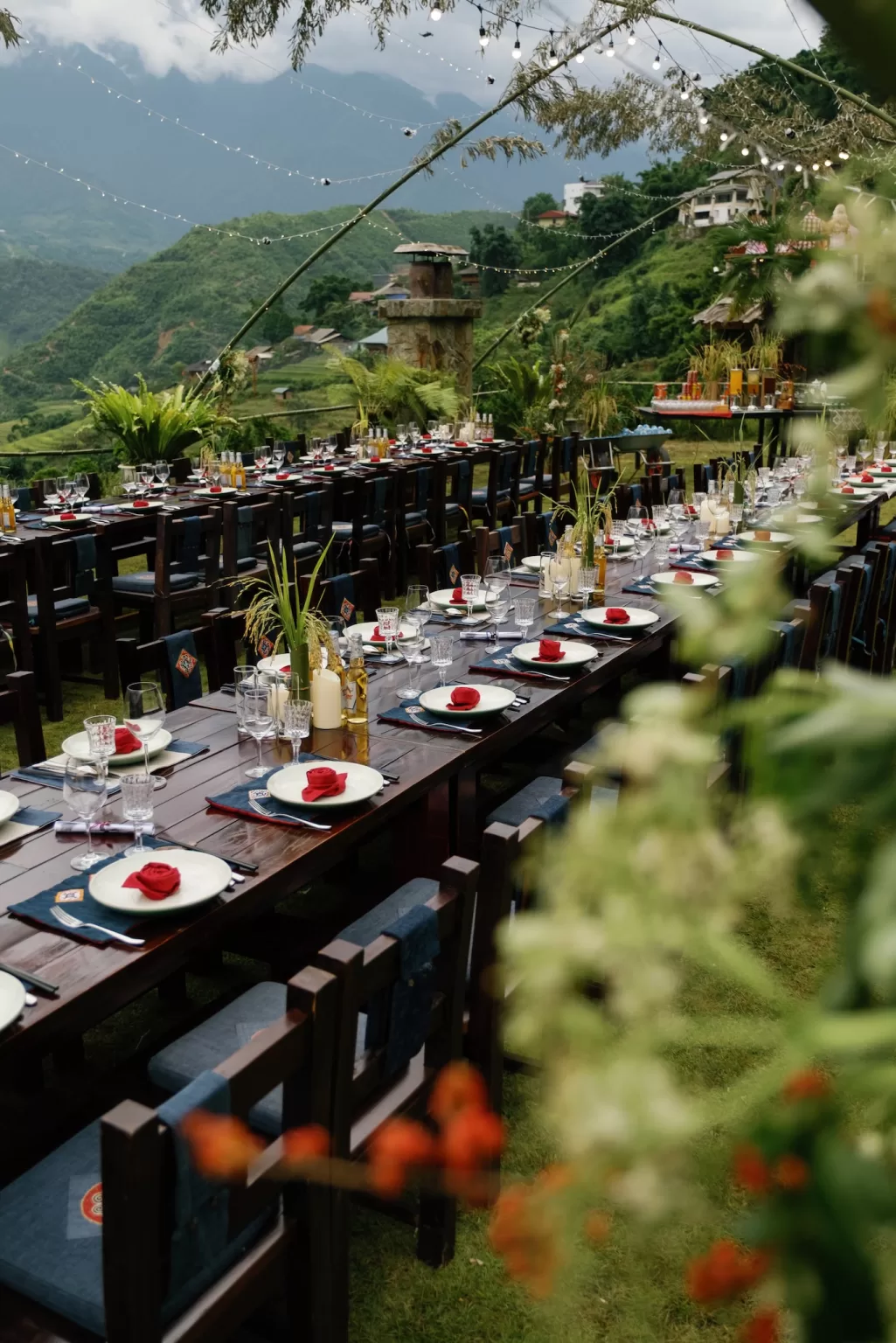 Long wooden banquet tables set outdoors at Ville de Mont Mountain Resort, overlooking layered green mountains beneath soft string lights.