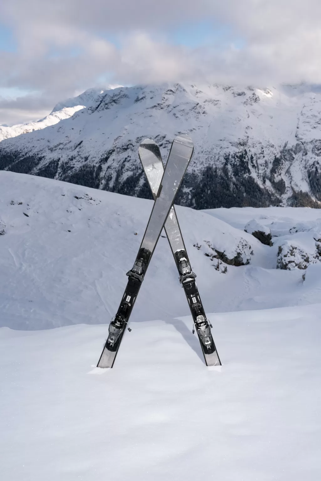 Pair of skis standing crossed in fresh snow with panoramic alpine mountains in the background.