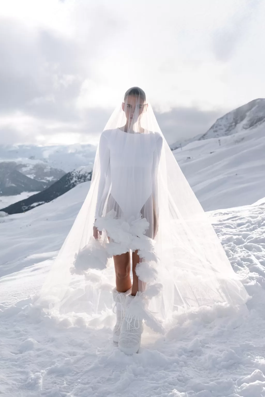 Bride wearing a long veil and winter boots standing in deep snow during an alpine bridal fashion shoot.
