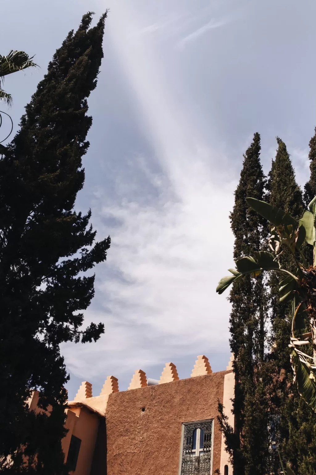 Tall cypress trees and terracotta walls of Beldi Country Club against a soft blue sky.