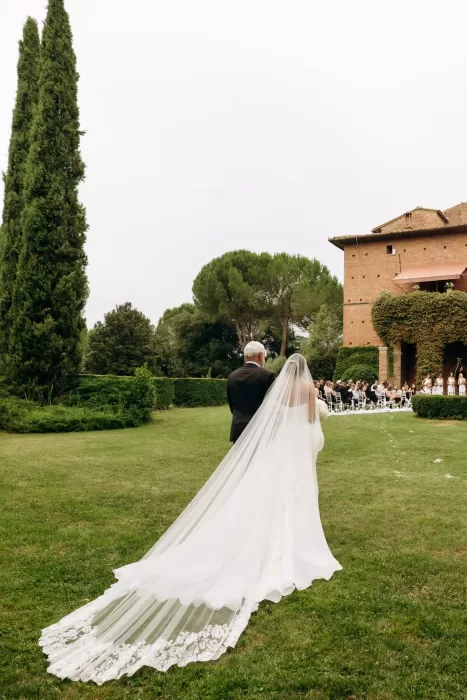 Bride walking toward outdoor ceremony with long veil flowing across garden at Castello di San Fabiano