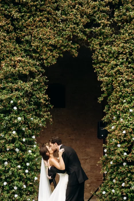 Bride and groom kissing beneath ivy-covered archway at Castello di San Fabiano