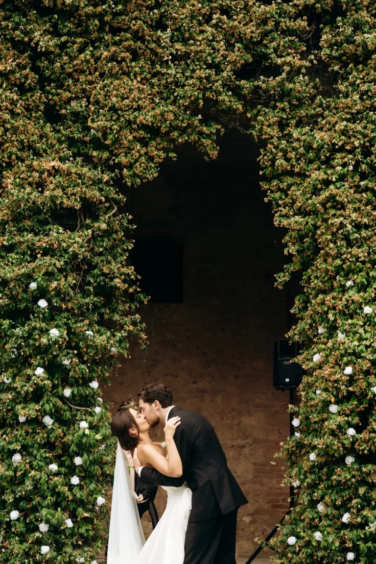 Bride and groom kissing beneath ivy-covered archway at Castello di San Fabiano