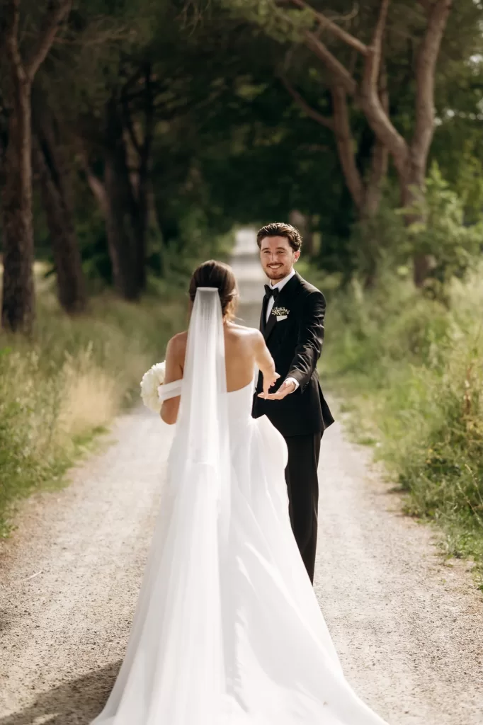 Groom reaching for bride during first look on tree-lined path at Castello di San Fabiano